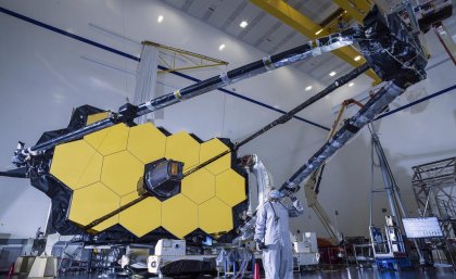 In order to detect the most distant and oldest galaxies, the telescope needs to be huge and kept extremely cold. Image: NASA/Chris Gunn. A person in a hazmat suit standing beneath a large telescope.